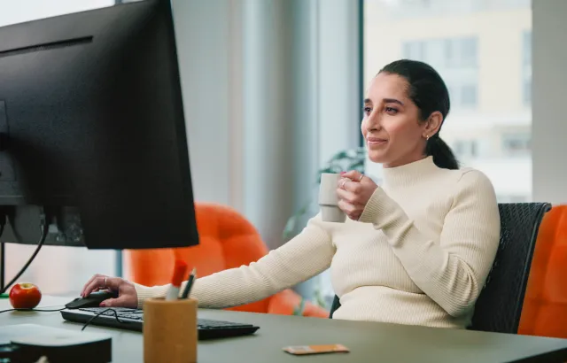 Woman happily working in an office behind her computer, with coffee in hand